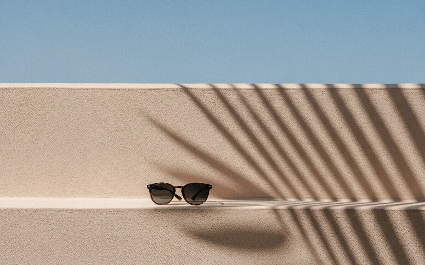 Luz tropical intensa y sombras de palmera sobre un muro claro, con lentes de sol